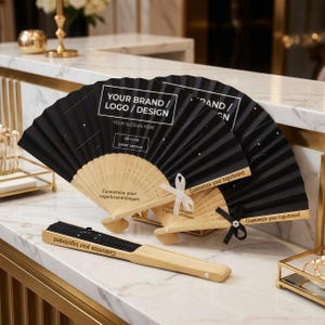 Peut inclure: Éventails pliants noirs et beiges avec le texte "YOUR BRAND / LOGO / DESIGN" et "Customize your logo/brand/slogan". Les éventails ont un design noir avec des étoiles blanches et sont attachés avec des rubans blancs et noirs. Les éventails sont en bois et en papier.