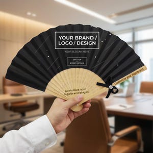 Pode incluir: Um leque dobrável preto com cabo de madeira, aberto. O leque tem o texto "YOUR BRAND / LOGO / DESIGN", "YOUR SLOGAN HERE", "OR CODE EVENT DETAILS" e "Customize your logo/brand/slogan". Uma fita preta com uma flor branca está presa.