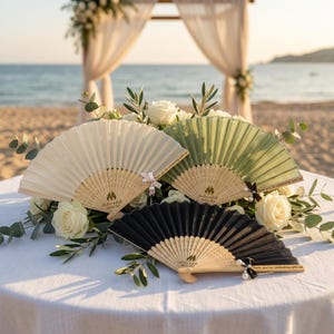 Peut inclure: Trois éventails pliants crème, vert sauge et noir, disposés sur une table avec des roses blanches et de la verdure. Chaque éventail a un manche en bois et un motif imprimé. Les éventails sont sur une table avec une plage et une arche de mariage en arrière-plan.