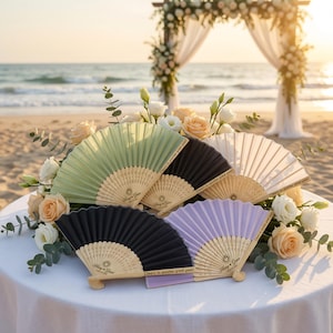 May include: Several open hand fans in shades of green, black, white, and lilac, displayed on a table with flowers. Each fan has a wooden handle and the text "Cheers to another great year!" The background shows a beach and a decorative arch.
