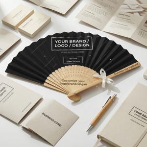 Op de afbeelding: Een zwarte vouwventilator met een houten handvat en een wit lint. De ventilator heeft de tekst "YOUR BRAND / LOGO / DESIGN" en "YOUR SLOGAN HERE". Visitekaartjes, brochures en een pen staan ook in de afbeelding.