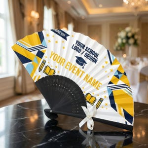 Puede incluir: Un abanico plegable decorativo con un mango negro y un abanico blanco y estampado. El abanico presenta diseños geométricos amarillos, azules y blancos, estrellas y el texto "YOUR EVENT NAME" y "YOUR SCHOOL LOGO / DESIGN". Una cinta blanca está atada en la base.