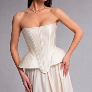 May include: A strapless cream-coloured corset top featuring vertical seam detailing and a structured peplum waist, paired with a matching cream-coloured skirt. The garment has a smooth, matte finish and a fitted bodice that flares at the hips.