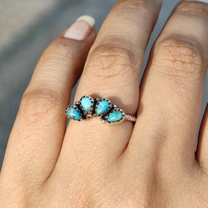 May include: A silver ring featuring five turquoise-colored stones arranged in a fan shape. The ring is set on a hand, showcasing the intricate details of the jewelry. The stones are a vibrant blue-green color.