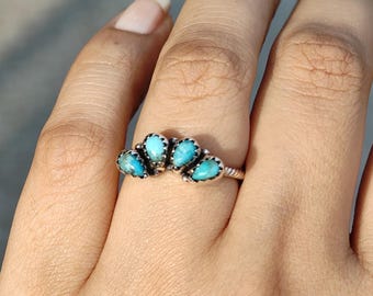 May include: A silver ring featuring five turquoise-colored stones arranged in a fan shape. The ring is set on a hand, showcasing the intricate details of the jewelry. The stones are a vibrant blue-green color.