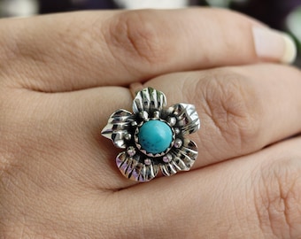 May include: A silver-colored ring featuring a flower design with detailed petals. The center of the flower holds a round, turquoise-colored stone. The ring is worn on a finger, showcasing the jewelry's design and color.