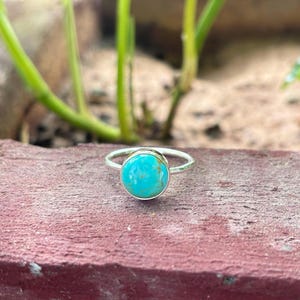 May include: A turquoise gemstone ring with a silver band. The ring is set on a red-brown surface.