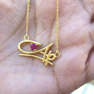 Può includere: Una collana d'oro con un ciondolo a forma di lettera stilizzata con una pietra preziosa di rubino.