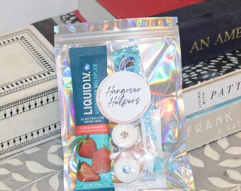 May include: A clear, holographic zip bag containing hangover recovery items. Includes a strawberry electrolyte drink mix, mints, a toothbrush, and a sticker that reads "Hangover Helpers". The bag is displayed on a patterned surface.