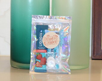 May include: A clear plastic bag containing small items, including a packet of Liquid I.V. Strawberry electrolyte drink mix. The bag also features a round sticker with the text "Bride's Babes". Two green glass bottles are in the background.