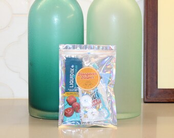May include: A clear, iridescent pouch labelled "Hangover Helper" contains various items. A blue packet of "Liquid I.V." is visible, along with other small items. Two green glass bottles are in the background.