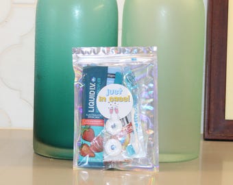 May include: A clear, iridescent pouch containing electrolyte drink mix, mints, and other items, with the text "Just in case!" and "Liquid I.V." on the packaging. Two large green glass bottles are in the background.