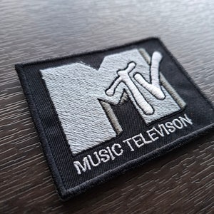 May include: A black and white embroidered patch featuring the MTV logo and the text "MUSIC TELEVISION".
