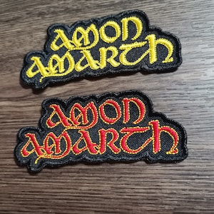 Puede incluir: Dos parches bordados con el nombre de la banda "Amon Amarth" en negro con letras amarillas y rojas. El parche superior tiene letras amarillas y el parche inferior tiene letras rojas.