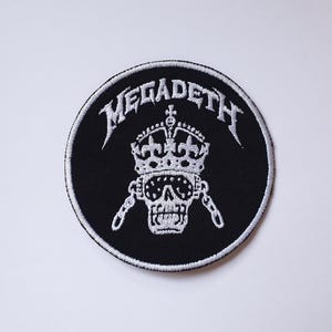 May include: Black and white embroidered patch featuring the band name "Megadeth" in a white cursive font. A white skull with a crown is in the centre of the patch.