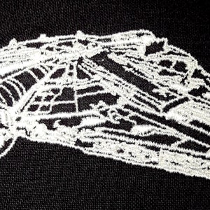 Könnte beinhalten: Weiß gesticktes Bild des Raumschiffs Millennium Falcon auf einem schwarzen Stoffhintergrund.