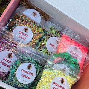 Peut inclure: Une boîte rose remplie de sachets de paillettes transparents. Les sacs sont étiquetés "Snake Skin", "Star Struck", "Dragon Tales", "Pineapple Express", "Fire" et "Drama Queen". Les couleurs des paillettes comprennent l'or, le vert, le rose et le jaune.