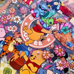 May include: A collection of colorful stickers featuring cartoon characters, flowers, and a Starbucks logo. The stickers include Winnie the Pooh, Stitch, and Tigger, with a variety of floral designs in pink, orange, and white. The background is a clear, textured surface.
