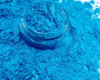 Puede incluir: Un pequeño frasco transparente lleno de polvo azul vibrante, rodeado por una generosa dispersión del mismo polvo. El polvo tiene una apariencia fina y texturizada, creando un contraste visual llamativo contra el fondo blanco.