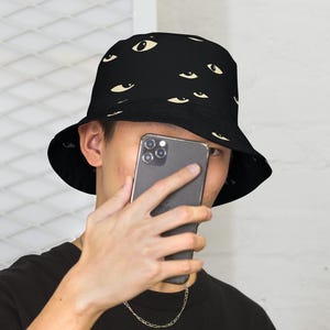 May include: Black bucket hat with an all-over eye pattern in cream. The hat is worn by a person wearing a black t-shirt and a silver chain necklace. The person is holding a smartphone.