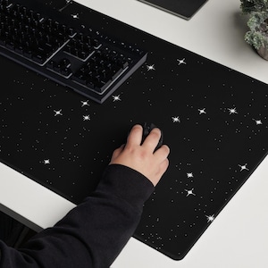 Puede incluir: Una alfombrilla de escritorio negra con un patrón de estrellas blancas, un teclado negro y un ratón de ordenador. La alfombrilla está sobre un escritorio blanco. Un monitor de ordenador está en el fondo.