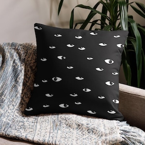May include: Black throw pillow with a white eye pattern. The pillow is on a couch with a blue and gray throw blanket.