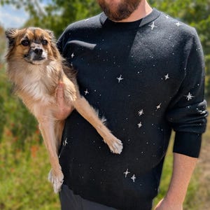 May include: A black long-sleeve jumper featuring a white embroidered star pattern across the front and sleeves. The knit garment has a crew neckline and ribbed cuffs, showcasing a celestial design with various sizes of four-pointed stars.