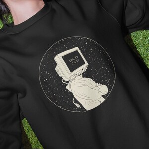 Könnte beinhalten: Schwarzes Sweatshirt mit Rundhalsausschnitt und einem kreisförmigen Grafikdesign einer Person mit einem Computermonitor als Kopf, mit dem Text "SPACE CADET". Das Sweatshirt ist auf einer Person zu sehen, die auf grünem Rasen liegt, umgeben von Schallplatten.