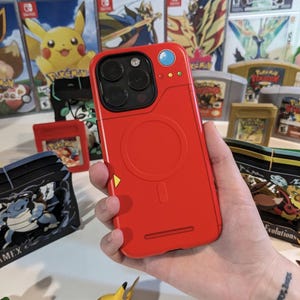 May include: A red iPhone case with a Poké Ball design. The case has a raised circle in the center and a yellow triangle on the bottom. The case is surrounded by various Pokémon video game cartridges.