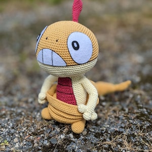 May include: A crocheted plush toy of a yellow and red Pokemon character with a white eye and a toothy grin. The toy is sitting on a bed of gravel.