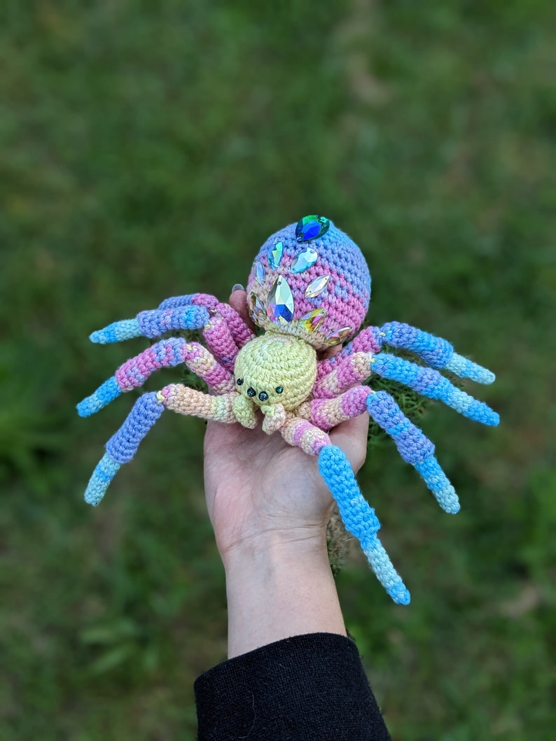This photo shows a crocheted spider that is rainbow in colour but looks like a black widow spider.It is being held in a palm of a hand against a grass background.