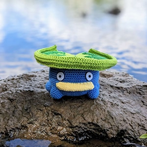 Puede incluir: Un juguete de peluche azul y verde tejido a crochet que se asemeja a un personaje de Pokemon. El juguete tiene un gran sombrero verde en forma de hoja y grandes ojos azules.