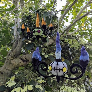 Może przedstawiać: Dwa ręcznie robione szydełkowe żyrandole wiszące na gałęzi drzewa. Żyrandole mają czarne, niebieskie, żółte i zielone akcenty. Te fantazyjne szydełkowe żyrandole są dekoracyjne i dodadzą niepowtarzalnego charakteru każdej przestrzeni.