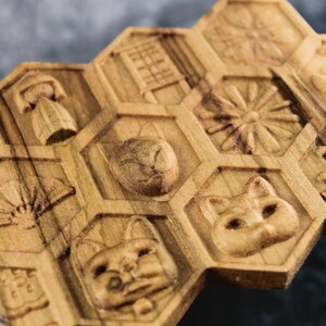 Puede incluir: Un azulejo hexagonal de madera con varias imágenes talladas, incluyendo un gato, un zorro, una flor y una abeja. El azulejo está hecho de madera marrón claro y tiene un acabado natural.