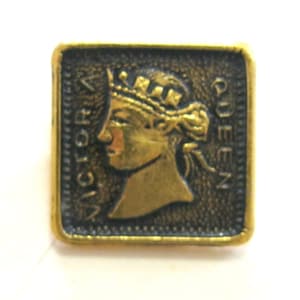 May include: A gold-colored metal button with a square shape and a raised image of Queen Victoria wearing a crown. The button has the words "VICTORIA QUEEN" inscribed around the edge.