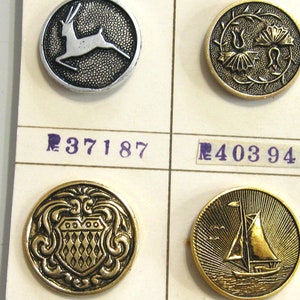 May include: Four round metal buttons with different designs. The top left button features a silver deer, the top right button features a gold floral design, the bottom left button features a gold shield with a diamond pattern, and the bottom right button features a gold sailboat.