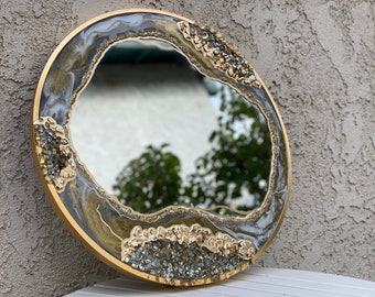 May include: A round mirror with a gold frame and a unique, abstract design. The mirror has a clear reflection and is surrounded by a layer of resin with gold and silver accents.