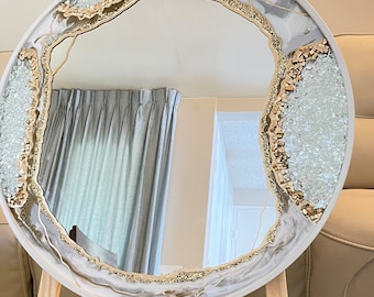 May include: A round mirror with a white and gold resin frame. The frame has a unique agate design with gold glitter and clear crystals. The mirror is on a wooden easel.