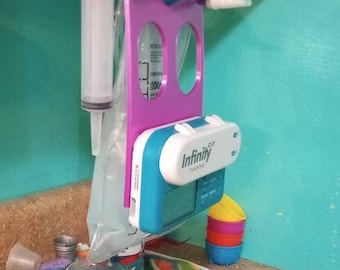 May include: A purple and blue medical device with a clear bag and a syringe. The device is labeled "Infinity" and is hanging from a cabinet. There is a colorful towel on the counter below the device.