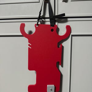 May include: A red plastic wall-mounted organizer with a black strap. The organizer has two hooks on the top and a rectangular shape with a cut-out on the bottom.