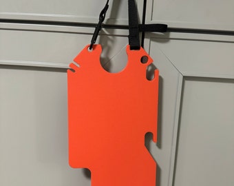 May include: A bright orange plastic safety device with a black strap. The device has a unique shape with cutouts and a hole in the center. It is hanging from a hook on a gray door.