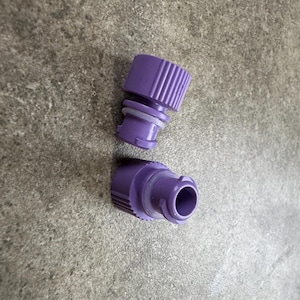 Peut inclure: Deux raccords de tuyau en plastique violets. L'un a un dessus nervuré et un joint circulaire. L'autre a une ouverture circulaire. Les deux raccords sont sur une surface grise texturée.