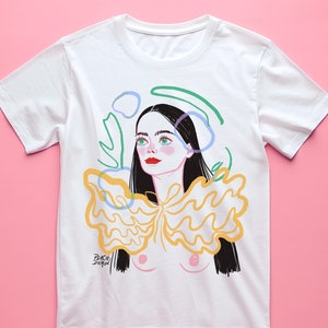 May include: White t-shirt featuring an illustration of a woman with black hair, red lips, and green eyes. The design includes a yellow and orange ruffled collar and abstract shapes. The text "PEACH DEMON" is printed on the shirt.