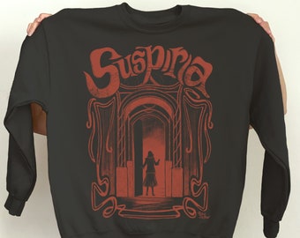 Könnte beinhalten: Schwarzes Sweatshirt mit einem roten Grafikdruck einer Frau, die in einem Durchgang steht, mit dem Wort "Suspiria" darüber. Die Grafik ist im Vintage-Stil.