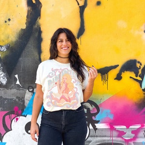 K&ouml;nnte beinhalten: Eine Frau tr&auml;gt ein wei&szlig;es T-Shirt mit einer Vintage-Illustration einer Frau, die eine Waffe h&auml;lt, und dem Text "Baby Doll" darauf. Sie steht vor einer gelben, pinken und blauen Graffiti-Wand.