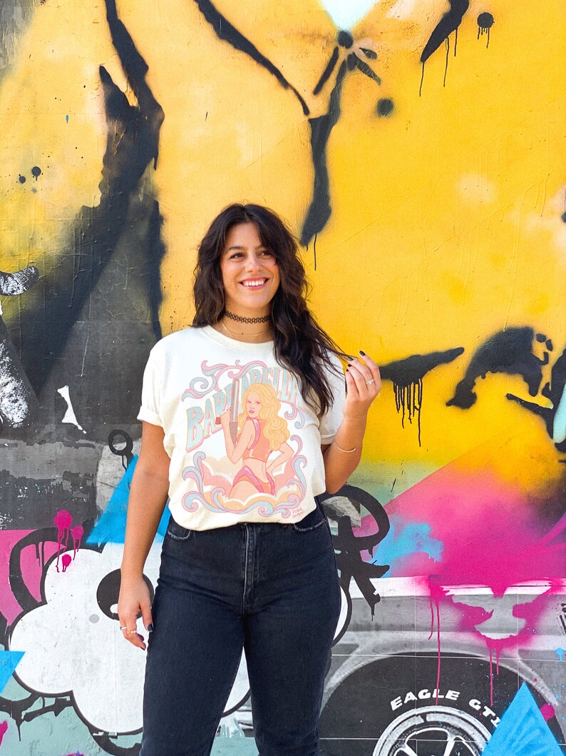 K&ouml;nnte beinhalten: Eine Frau tr&auml;gt ein wei&szlig;es T-Shirt mit einer Vintage-Illustration einer Frau, die eine Waffe h&auml;lt, und dem Text "Baby Doll" darauf. Sie steht vor einer gelben, pinken und blauen Graffiti-Wand.