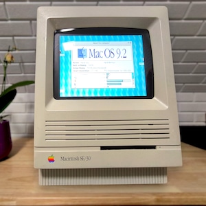 Könnte beinhalten: Ein Vintage Macintosh SE/30 Computer mit einem Bildschirm, der Mac OS 9.2 anzeigt. Der beige Computer hat einen eingebauten Bildschirm und einen Schlitz für eine Diskette. Links vom Computer steht ein violetter Blumentopf mit einer Pflanze.