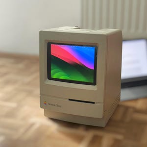 Op de afbeelding: Een beige Macintosh Classic computer met een vierkant scherm dat een kleurrijke abstracte achtergrond toont. Het voorpaneel bevat een sleuf voor een diskettestation en het Apple-logo met de tekst Macintosh Classic eronder.