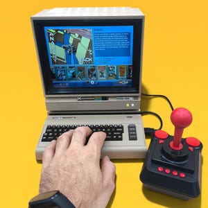 May include: A beige Commodore 64 computer with a screen displaying the game Anarchy, featuring a black keyboard and a black joystick with a red handle and buttons. The screen shows game text and a menu of various game titles at the bottom.