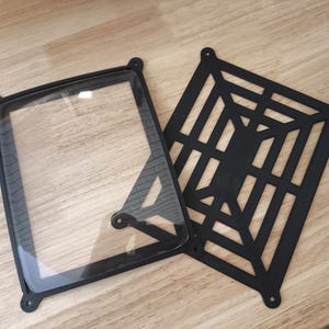 May include: Two black, square components. One is a frame with a clear, rectangular window. The other is a black, geometric, openwork panel. Both have rounded corners with mounting holes. They are on a light wood surface.